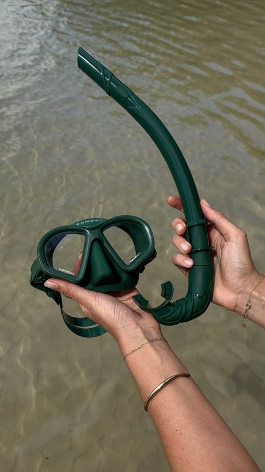 Hands holding a green snorkel mask and snorkel above clear shallow water.